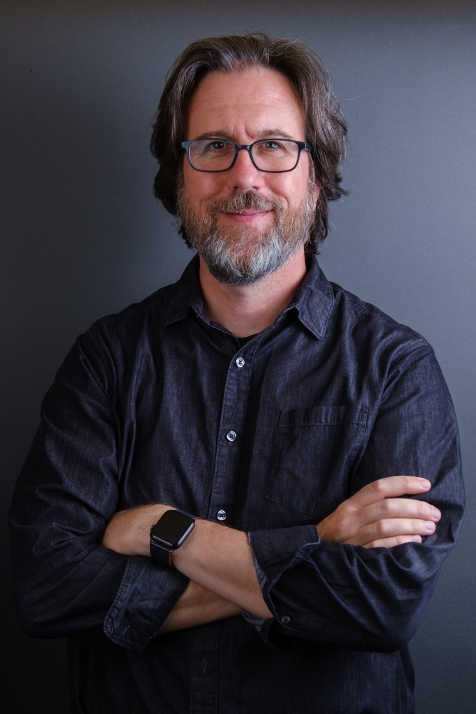Portrait of Jeff Carlson, a white man with dark hair, black glasses, and a dark and gray beard. He's wearing a dark gray button-down shirt with the sleeves rolled up. His arms are crossed and he's smiling at the camera.