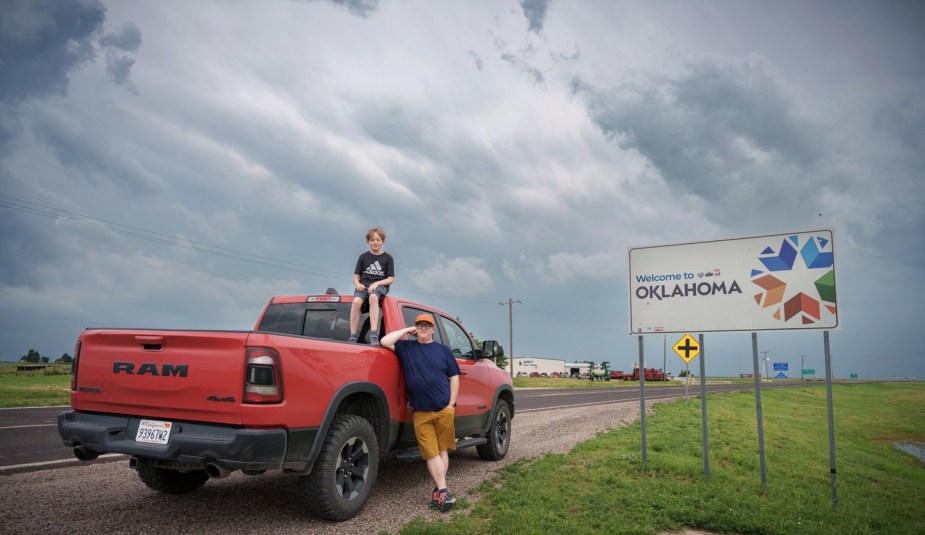 A father and son lean against a red pickup truck next to a sign reading "Welcome to Oklahoma"
