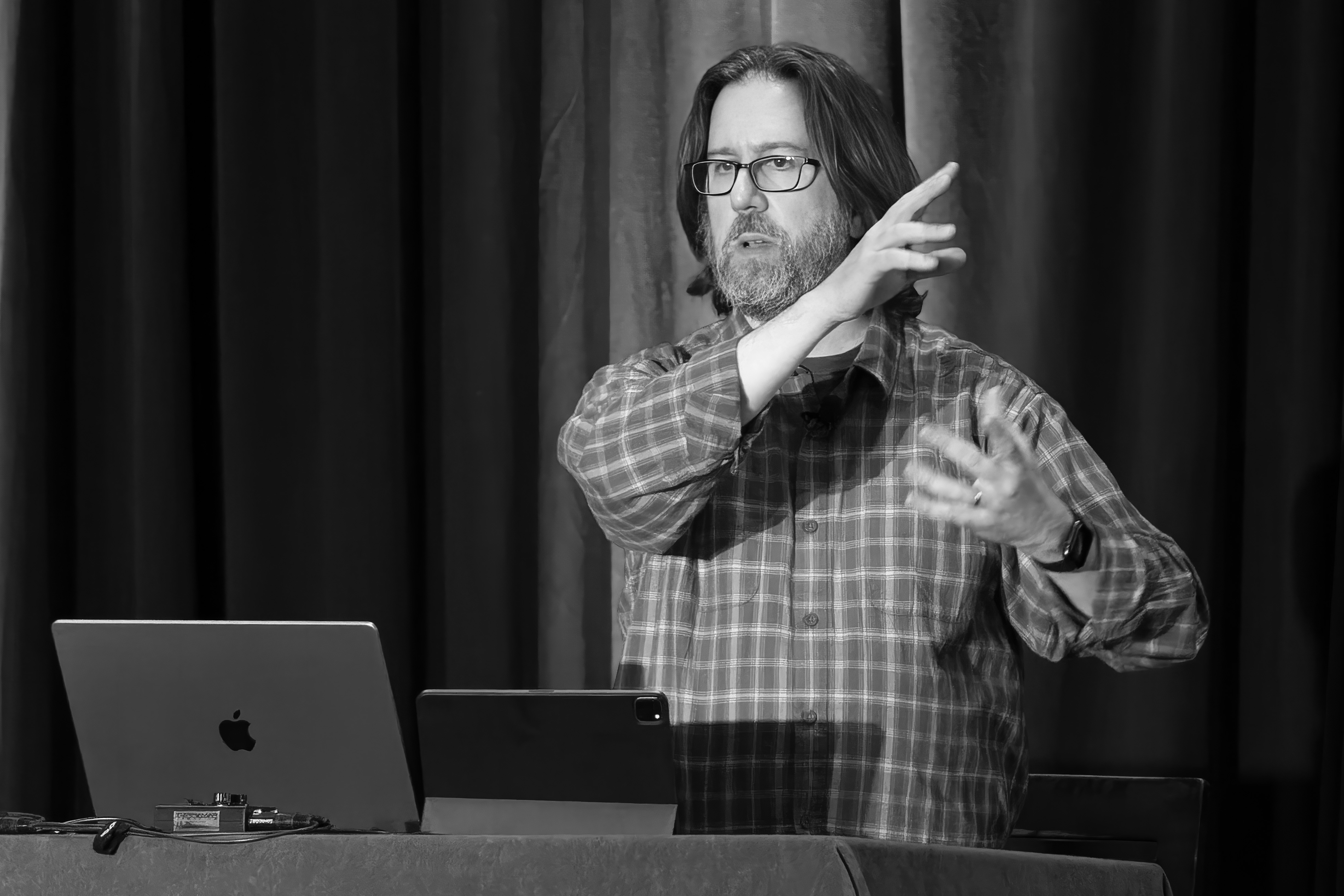 Black and white photo of Jeff Carlson, a man wearing a striped shirt, gesturing while speaking to an audience and standing in front of a table with a MacBook Pro and an iPad Pro.