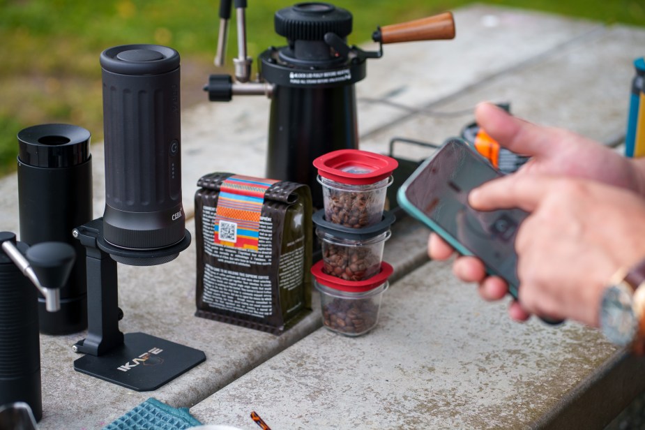 A person holds a smartphone and uses an app to set parameters for brewing espresso in a portable machine on a park bench.