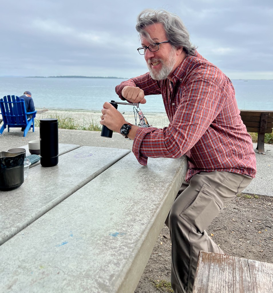 A man crouches near a park bench while hand-grinding coffee beans.