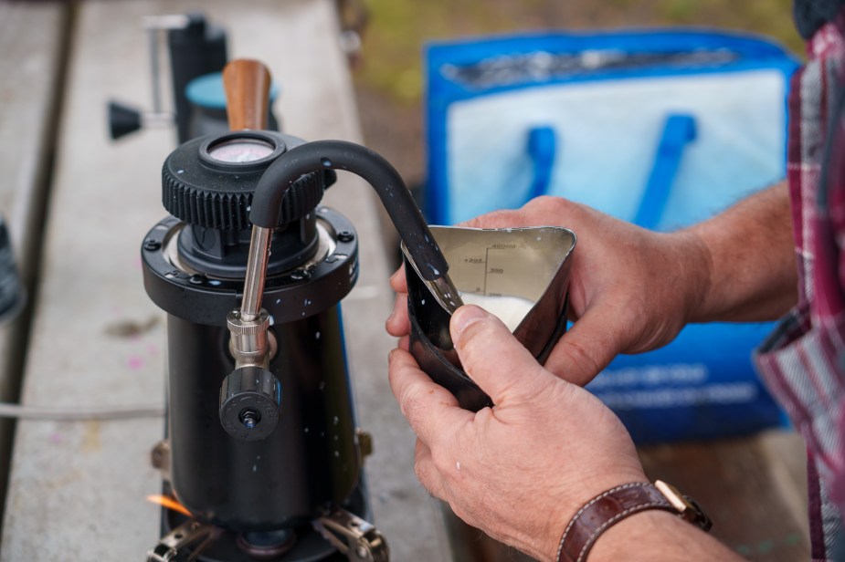 Closeup of a two hands holding a milk pitcher and steaming the milk using a portable steamer on a park bench.