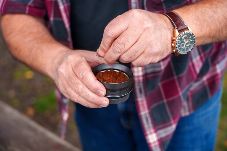 Closeup of a man stirring a basket of espresso grounds with a metal tool.